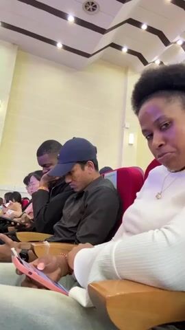 The video shows a woman in the foreground, to the right, looking at the camera with a slight smirk. She is wearing a white, ribbed sweater and a necklace. She is holding a pink smartphone in her right hand, and her left hand is resting on her lap. Behind her, seated in a theater-like setting, are several other people. To the left of the woman in the foreground, a man in a dark shirt and a baseball cap is looking down at his phone. To his left, another man in a black shirt is also looking down, possibly at his phone. Further to the left, a woman with glasses is visible, also looking down. The seats are upholstered in red and wood. The lighting appears to be from overhead fixtures on a light-colored ceiling. The overall impression is of an audience waiting for an event to begin or during a break.