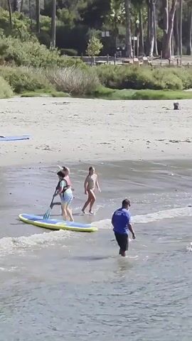 This video captures a vibrant beach scene in Dana Point, California, bathed in the bright light of a sunny day. The camera focuses on the shoreline where several individuals are engaged in water activities. In the foreground, the clear, shallow water laps at the sandy beach.

The primary subjects are a group of people, including children and adults, interacting with paddleboards. A young girl in a blue life vest is seated on a yellow paddleboard, paddling with a dark-colored oar. An adult male, wearing a blue t-shirt and black shorts, stands in the water near the paddleboard, appearing to offer assistance or guidance.

Further towards the shore, two young girls in swimsuits are playing in the shallow water. One girl, with blonde hair, is running with her arms raised, while the other, also with blonde hair, walks towards the water's edge. Another young girl in a pink bikini is also visible near the water.

On the sandy beach in the background, there's a hint of a park or recreational area with tall palm trees and greenery. A few other individuals can be seen on the beach, including someone in a red shirt and shorts walking away from the water, and another person in the distance. The overall atmosphere appears to be one of casual recreation and enjoyment on a pleasant day. The time of day is clearly daytime, likely late morning or early afternoon, given the strong sunlight and clear skies. There is no visible text in the video.