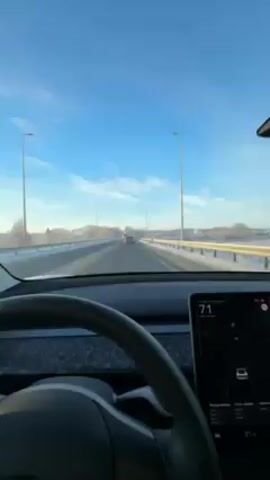 The video shows a view from inside a car, likely a Tesla, driving on a highway. The dashboard is visible, displaying a map and a speed of around 70 km/h. The landscape outside is snowy, with a clear blue sky above. Other cars are visible on the highway. The camera pans slightly, showing the car's interior, including the rearview mirror with an air freshener hanging from it.