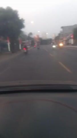 The video shows a blurred, first-person view from inside a vehicle, likely moving. The scene is a road with buildings and trees lining one side. Several motorcycles are visible on the road, some with their lights on. Streetlights are also present. The sky is overcast, suggesting it is either early morning or late evening. The motion blur indicates the vehicle is in transit.