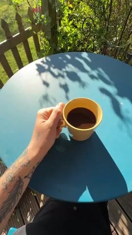 A person is sitting at a blue round table in a garden. The camera is positioned above the table looking down. Initially, the person’s hand with a tattoo is holding a yellow mug filled with a dark liquid, likely coffee. The hand lifts the mug, moves it away from the table, and then returns it to the table. The table is on a wooden deck, and behind the table is a green bush and a white wooden fence. The sun is shining, creating shadows on the table.