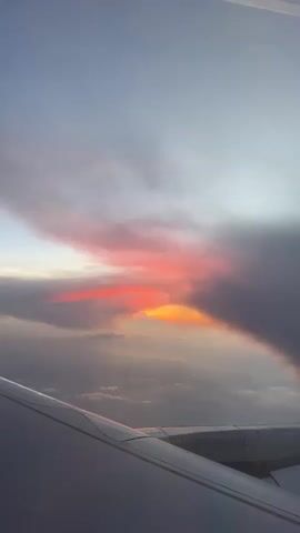 This is a vertical, POV shot from an airplane window. In the foreground, a portion of the airplane's wing is visible, extending from the bottom left corner and curving towards the center of the frame. The wing is white with some faint gray shadowing.

The background shows a vast sky with clouds. The sky transitions from a light blue or gray at the top to a lighter, hazy horizon. A dramatic sunset or sunrise is occurring, with vibrant colors painting the clouds. A large band of dark gray clouds dominates the right side of the frame, partially obscuring the colorful sky. In the gap between the clouds, the sky is illuminated with brilliant shades of orange and intense red, suggesting the sun is low on the horizon. Streaks of lighter clouds are visible below the colorful band, creating a layered effect. A faint, thin contrail is visible in the upper right corner of the frame, indicating the plane's movement through the atmosphere. The overall impression is one of a breathtaking aerial view of atmospheric phenomena.