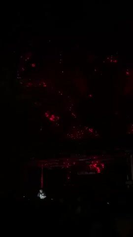 A person is visible in the foreground against a dark background, appearing to be on a stage or platform. They are dressed in light-colored clothing. Above them, a stage rigging with red lights is illuminated, casting a red glow on the ceiling or upper structure of the venue, creating a scattered, fiery effect with red lights. The atmosphere is dark, indicating an evening or indoor event, likely a performance or concert, with the red lighting being a key visual element.