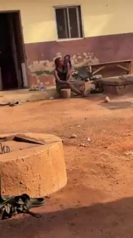 A woman is sitting on a tree stump, holding a young child. She is wearing a black tank top and a patterned skirt. The child has blue hair and is wearing nothing. The woman is kissing the child on the lips. They are sitting in a dirt yard, with a building in the background. There is a water bottle on the ground nearby.