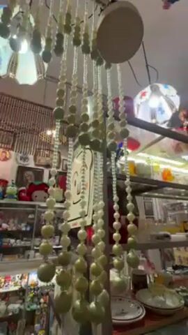 The video shows a cluttered shop interior. Hanging from the ceiling is a decoration made of strings of white and green beads, with a white disc-shaped lamp partially visible above it. Behind the beads, several shelves display a variety of items, including Hello Kitty merchandise, plates, figurines, and framed pictures. Two additional decorative lamps with colorful floral patterns hang from the ceiling. The overall environment appears busy and eclectic, filled with a diverse collection of goods.