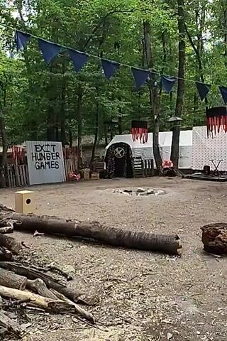 The video displays an outdoor scene, possibly a campsite or event space, set within a wooded area. A large log lies horizontally in the foreground. In the middle ground, a large, crudely painted sign reads "EXIT HUNGER GAMES." Behind the sign, several tents or structures are visible, adorned with triangular blue and red banners. A central tent has an ornate, circular design above its entrance. The ground is covered in dirt and wood chips, with more logs and branches scattered around. The overall impression is that of a themed gathering or immersive experience. There are no people visible in the video.