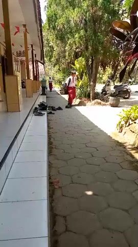 The video shows an outdoor walkway next to a school building. Several pairs of shoes are lined up along the base of the building. Two young students in school uniforms are walking down the path away from the camera. One student is carrying a backpack. A scooter is parked to the right of the path. Trees and foliage are visible in the background.