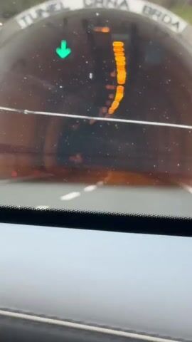 The video shows a view from inside a vehicle traveling through a tunnel. The tunnel walls are illuminated by rows of yellow lights, creating a warm, orange glow. The road ahead is marked with white lines, indicating lanes. Dust or water particles are visible on the windshield. A green directional arrow is briefly visible in the upper left corner of the frame. The camera perspective suggests the video is recorded from the driver's or passenger's seat.