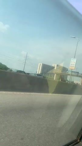 The video captures a view from inside a vehicle moving along a road. A large concrete barrier is prominent in the foreground. Beyond the barrier, a pedestrian bridge spans across the road. Several people are visible walking on the bridge. In the background, a multi-story building with many windows is situated behind the bridge. Two large billboards, one on each side of the pedestrian bridge, display advertisements for "Elite City." The sky is blue with scattered clouds, and streetlights are visible along the road and the bridge. Power lines and poles are also present in the distance. The overall environment appears to be an urban setting with elevated infrastructure.