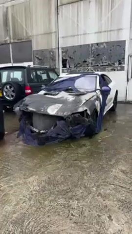 The video shows a white car that has been badly burned.  The hood and front end are significantly damaged and covered in soot and ash. A blue cloth is draped over the car, partially obscuring the damage. Behind the burned car is a black SUV. The scene is set outdoors on a wet, concrete surface in front of a large, corrugated metal building.