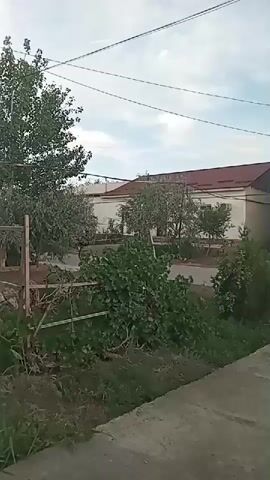 The video shows a person walking on a paved path. The person is not visible in the frame, but their movement suggests they are walking away from the camera. The path is surrounded by greenery and a building can be seen in the background. The sky is overcast.