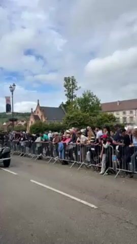 The video shows a black and red vintage car driving on a street lined with a crowd of people behind a metal fence. The car passes the crowd at a slow pace. The driver is visible, as is another person in the passenger seat. The background shows buildings and trees under a cloudy sky.