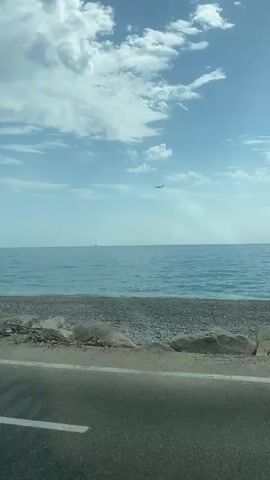 The video shows a wide shot of a beach and the ocean. The sky is blue with some white clouds. An airplane is visible flying in the distance. In the foreground, there is a rocky shore with some large rocks. There is also a paved road or path in the lower part of the frame. The ocean is calm and a few small waves are visible. A person is partially submerged in the water. A blue umbrella is set up on the beach.