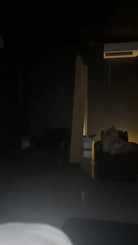 The video shows a dimly lit room with two dark armchairs positioned on either side of a central area. A person is reclined in the right armchair, looking at a phone, and covered with a light-colored towel or sheet.  Light emanates from behind the chair, casting a soft glow on the wall. Cream-colored curtains hang on either side of the chair, adding to the sense of privacy.  An air conditioning unit is mounted high on the wall above the chair. The floor appears to be tiled. The overall ambiance is tranquil and private.