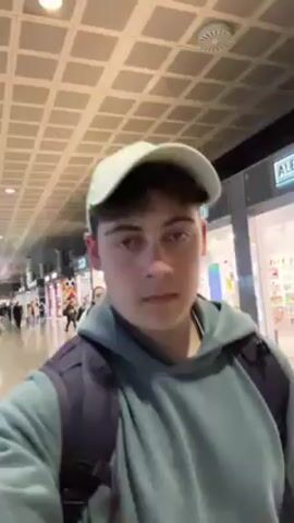 A young man is holding a camera, recording a selfie-style video in an airport. He is wearing a light green hoodie, a dark backpack, and a light-colored baseball cap. Behind him, a young woman with long hair is pulling a teal suitcase and walking in the same direction. The airport has a high, tiled ceiling. Shops are visible on the right side of the hallway, while a seating area with many people is on the left. The floor is a light color, reflecting the overhead lights. Other people are walking through the airport in both directions.