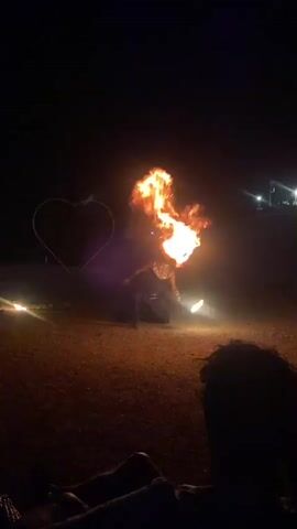 A person kneels on the ground, surrounded by darkness, with a bright flash of fire erupting from their head. To the left, a large, lit heart shape is visible against the dark sky. The ground is covered in gravel. In the foreground, the back of a person's head and shoulders are visible, facing away from the action. The video captures the fiery display for a few seconds, with the flames pulsing and shifting.