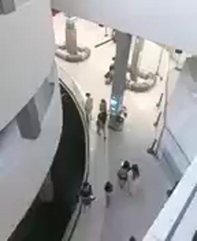 This media file is a video depicting an interior scene, likely a modern public building such as a museum or shopping mall, based on the architectural style. The setting features clean, curved white walls and a bright, open atrium. Several people are visible in the space, walking and interacting. In the upper portion of the frame, a group of individuals can be seen walking together. Further down, another group of people, including what appears to be a family with children, are gathered. The lighting suggests it is daytime, and there are no indicators of weather conditions. The overall atmosphere appears casual and relaxed. There is no visible text within the frame. The camera angle is from a high vantage point, looking down into the atrium, providing a broad overview of the activity below.