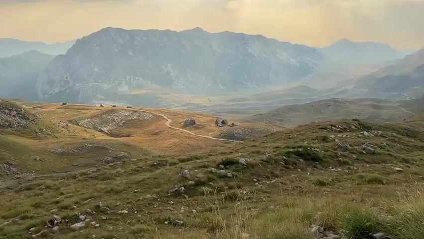 The video shows a panoramic view of a mountainous landscape with scattered rustic wooden cabins. The terrain is covered with dry, golden grass and rocks. A dirt road winds through the scene, leading towards the cabins. Several sheep are grazing or walking around the cabins. There are two cars parked near the cabins, one with its trunk open. In the background, the mountains are hazy, suggesting distance or atmospheric conditions. The lighting suggests it is either early morning or late afternoon, with a warm, soft glow over the landscape.