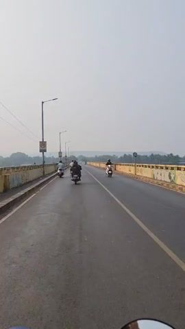 The video shows a view from a moving scooter driving across a bridge. The bridge has two lanes with a single yellow line dividing them. There are several other scooters and motorcycles on the bridge, moving in the same direction. Street lights line the bridge. The bridge has yellow concrete barriers on either side, marked with graffiti. In the distance, trees and a hazy sky are visible.