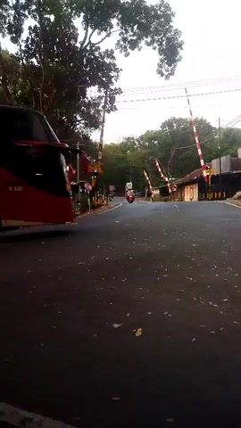 The video begins with a view of a road with a bus on the left side. The bus is red and black and has the number R 020 on it. The road is asphalt and has some debris on it. In the background, there are trees and power lines. There are also railroad crossing gates. One gate on the right side is down, indicating a train is approaching or passing. A person on a motorcycle is visible in the distance, approaching the crossing. The sky is overcast. The video is shaky, suggesting it was recorded while moving.