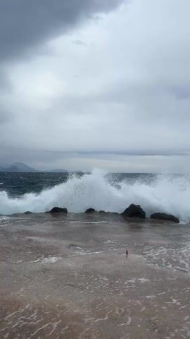 The video shows a choppy ocean on a cloudy, overcast day. Waves crash against a rocky shore, with white foam visible. The horizon line is visible in the distance. A cement surface is in the foreground, showing the rough edge of the pier with a red object laying on the ground.