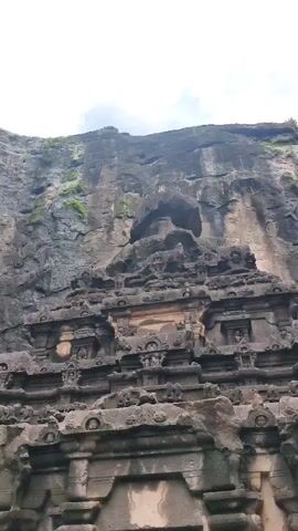 A low-angle video captures the exterior of a massive stone structure carved into a cliff face. The architecture is intricately detailed with multiple tiers of carvings, depicting figures and decorative motifs. The stone appears weathered and grey, with some areas showing signs of erosion. Vegetation, appearing as green moss or small plants, is visible growing on the upper parts of the cliff. The sky above is overcast with white clouds. The camera slowly pans upwards, revealing more of the colossal scale of the rock-cut structure. There are no people visible in the frame.