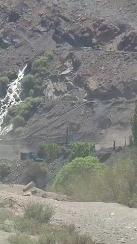The video shows a cascading waterfall flowing down a rocky mountainside in a remote area. The water is white and foamy, contrasting against the dark, gray rocks. Patches of green vegetation dot the hillside, and there are a few small structures at the base of the falls. Utility poles run along the lower edge of the slope, and there is a dirt road in the foreground. A truck can be seen near the base of the waterfall. The camera appears to be positioned from a moving vehicle.