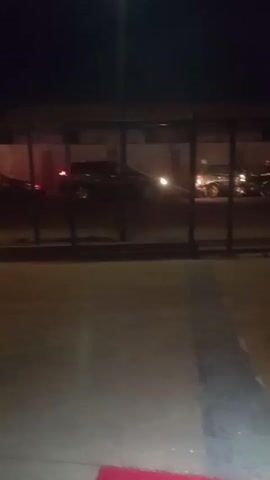 The video shows an outdoor scene at night. There are several parked cars visible behind a low metal fence. A few people are standing near the fence, though details are unclear due to the low lighting. A person in a dark jacket and pants walks towards the group of people. The surroundings appear to be paved, possibly a parking lot or a similar outdoor area.