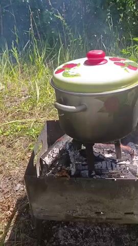 A large pot with a lid decorated with strawberries sits on a metal grill filled with glowing embers. A hand lifts the lid, revealing a pot of cooked crawfish, noodles, a bay leaf, and a halved onion. Steam rises from the pot as it sits outdoors in a grassy area.