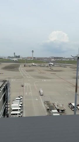The video is a static, slightly shaky, medium shot of an airport tarmac on a cloudy day. The foreground shows a dark gray ledge or balcony. Beyond this, the tarmac is visible, with several white vans parked in a row on the left. Further back, a series of baggage carts and a tow truck are parked. In the middle ground, an airplane is visible on the tarmac, appearing to be preparing for or just finishing a maneuver. There are other airplanes in the distance, as well as the characteristic control tower of an airport. The sky is overcast with a large, fluffy white cloud visible.