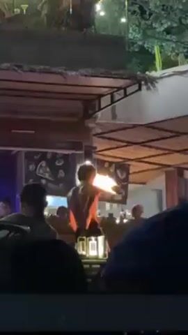 The video shows a fire dancer performing at an outdoor restaurant. The dancer, a shirtless male, skillfully manipulates flaming batons. The restaurant is decorated with string lights and has a thatched roof structure. Several patrons are seated at tables, watching the performance. The scene is lively with the sounds of music and cheering.