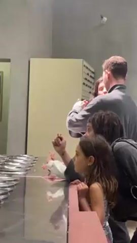 A group of six people, including four children, are standing in front of a large stainless steel counter in what appears to be a storage room. The counter is filled with rows of silver-colored metal discs. A man in a gray shirt stands at the back of the group, partially hugging a woman with long curly hair. Three children stand at the front, looking at the counter, one pointing towards the metal discs. The room has concrete walls and a light-colored cabinet on the side. A painting can be seen on the left wall. The lighting in the room is slightly dim.