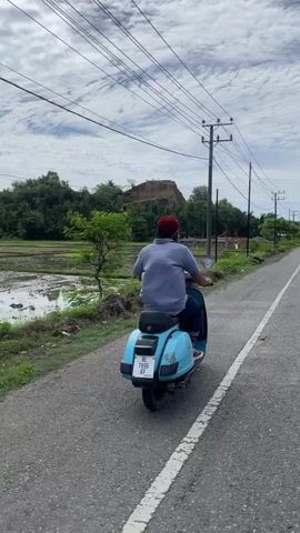 A person wearing a blue shirt and a red cap rides a light blue scooter on a paved road. The scooter has a white license plate with black text. The road is on the right side of the frame, with a white line marking the edge. To the left of the road is a patch of dirt and grass, followed by a flooded field. In the background, there are trees, a pile of dirt, and power lines supported by poles. The sky is cloudy.