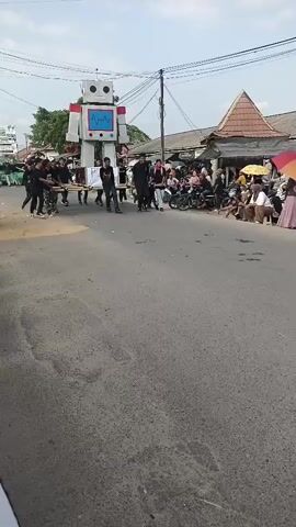 A group of young people dressed in black are carrying a large, light-colored robot costume on poles in a procession. The robot is tall and has a screen on its chest displaying a blue and red waveform. They are walking down a paved street lined with spectators and market stalls. Some of the spectators are seated on the side of the road, while others are standing. A person in the foreground is holding a colorful umbrella. The sky is overcast.