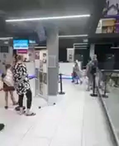 The video shows a group of people in what appears to be an indoor public space, possibly a transportation hub or a shopping area. The lighting suggests it could be daytime, but there are no clear indicators of natural light. The scene is bright with artificial lights, including a prominent strip light on the ceiling.

There are several individuals visible, mostly facing away from the camera or in profile. In the foreground, a person in a black and white patterned top and black leggings is standing, facing towards a set of displays or information boards. Another person in a green t-shirt and denim shorts is visible to the left, also facing away. Further into the scene, a queue of people is forming, with some individuals standing and others possibly moving. There are stanchions with black poles and red ropes creating a waiting line. In the background, there are counters or information desks, one of which has a blue light and a visible number "6" on it. There are also screens displaying images, one of which shows what looks like a landscape or sky.

The overall mood seems neutral; the people are engaged in everyday activities like waiting in line or browsing information. There are no obvious signs of distress or excitement. The environment appears modern and clean, with tiled floors and a dark ceiling. The location context provided indicates Reda, Poland, though no specific Polish text is visible in the frames.
