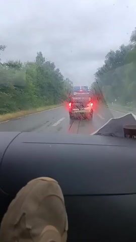 The video is shot from inside a vehicle, looking out through the windshield, which is wet with rain. In the foreground, a foot wearing a tan sneaker is visible resting on the dashboard. Ahead, on a wet highway, a dark-colored van with its brake lights illuminated is driving away. The van has a logo or text visible on its rear, though it is blurry. Trees line both sides of the highway, and the sky is overcast. Other vehicles are visible further down the road. The video is short, lasting only a few seconds.