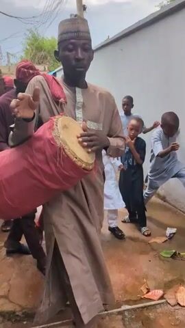 The video captures a lively outdoor procession in Unguwar Rimi, Nigeria, featuring several individuals dressed in traditional attire.

**Subjects:** The main subjects are several men and a few women forming a walking procession. Prominently featured are a man in a light brown traditional outfit and cap, playing a large talking drum wrapped in red fabric, and another man in a matching red tunic, trousers, and cap, playing a smaller drum with a stick. A young child in a dark blue outfit runs alongside the man in red. Other participants include men in maroon and cream-colored traditional wear, some wearing caps, and women dressed in hijabs and patterned outfits.

**Objects:** The central objects are two different types of drums being played, traditional caps (fila), various traditional garments, a long wooden staff held by one man, and a metal tray briefly visible in the foreground.

**Scene:** The scene is an outdoor paved pathway or alleyway, likely within a residential area. Buildings with light-colored walls line the path. Overhead, power lines and a utility pole are visible against an overcast sky.

**Activity or event taking place:** The group is engaged in a vibrant procession, possibly a cultural or celebratory event such as a wedding, naming ceremony, or festival. The men are actively playing drums and walking, setting a rhythmic and festive tone. Some participants, especially the man in red, display animated movements, dancing and moving to the drumbeats.

**Notable details:** The time of day is daylight, indicated by the natural lighting, though the sky is overcast, suggesting a cloudy day. The drumming is a focal point, with the musicians actively performing as they move. The overall mood appears celebratory and communal, with participants moving together to the rhythm of the music.

**Visible text:** There is no discernible text visible in the video.