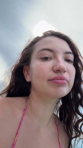 A woman with brown hair in a pink bikini is on a boat. She looks into the camera, smiling, and brushes her hair out of her face. The sea and sky are visible in the background. Other people are on the boat behind her.