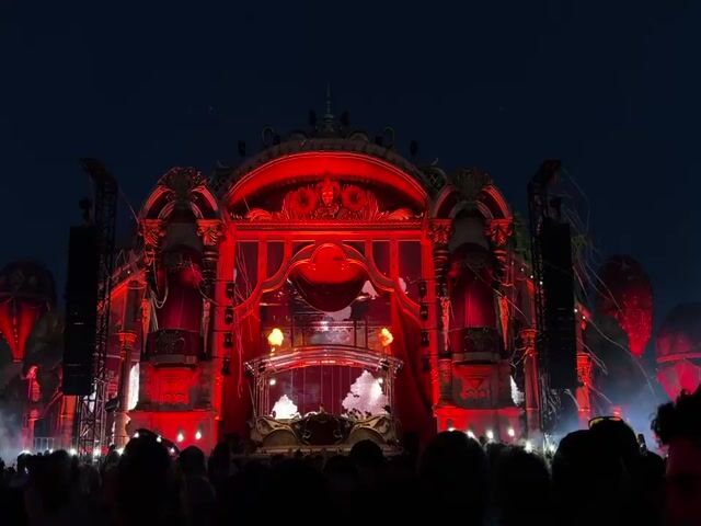 The video displays a nighttime outdoor concert or festival with a large, elaborate stage. The stage features a grand, ornate structure with red and gold accents, resembling a baroque or fantasy palace. Flames erupt from the sides of the stage, and various lights illuminate the area. A crowd of people is visible in the foreground, their backs to the camera, facing the stage. The sky is dark, indicating it is night. The overall atmosphere appears to be one of excitement and spectacle.