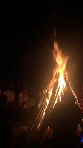 A bonfire burns brightly in a dark outdoor setting. Several people are gathered around the fire, some of whom are playing drums. The fire is tall and made of logs, with sparks flying upwards. The people are silhouetted against the firelight. The scene appears to be a gathering or celebration.