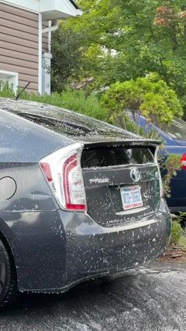 The video shows the rear of a dark gray Toyota Prius being washed. The car is covered in soap suds. A blue and white washing brush is visible on the left side of the frame, with a hand wearing a blue glove holding it. The car is parked on a paved surface, likely a driveway, with a house visible in the background. Green trees and bushes are also in the background. The license plate on the car reads VCD-1662.