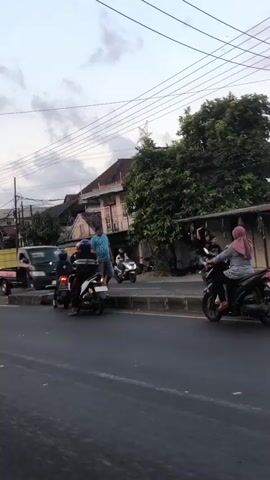 A low-angle, medium shot captures a street scene with several motorcycles and a few people. The sky is overcast with clouds. On the left, two motorcycles are stopped, one with two people on it, and another is further back. In the center, a man in a blue shirt and shorts stands near a white motorcycle, talking to two people on the motorcycle. To the right, a white motorcycle with a person on it is stopped, and another motorcycle with a person is further ahead. In the background, there are buildings, trees, and power lines. The road is paved. The overall impression is a busy street with motorcycles in motion.