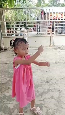 A young child, wearing a pink dress and sandals, dances on a concrete surface in front of a gate. The child has dark hair and is smiling, moving their arms in a rhythmic fashion. The gate is made of white metal bars, with a building visible in the background. There are trees and foliage visible overhead, suggesting an outdoor setting. The ground is a light gray concrete.