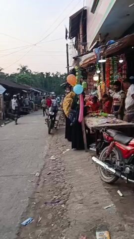 The video shows a street scene in an outdoor market area. A man wearing a patterned shirt rolls up his sleeve and looks at his watch while standing on the street. People are seen walking and standing in the market area. There are small shops with corrugated metal roofs along the street. A motorcycle is parked. A man wearing a helmet gets on the motorcycle and looks at his phone. There are also balloons attached to the side of a shop.