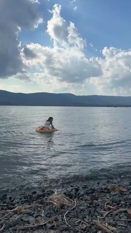 This is a video clip taken from a shoreline, looking out onto a large body of water, likely a lake. The water is calm, with gentle ripples. The shore is made up of pebbles and some dried vegetation. In the distance, rolling hills covered in trees form a backdrop against a bright sky filled with scattered, fluffy white clouds. The sun is casting a warm light, suggesting it is either morning or late afternoon.

The main subject is a young girl, who appears to be of Asian descent, sitting on a colorful inflatable ring in the water. She is wearing a swimsuit with a rainbow-like pattern. She is reaching out with her hands, seemingly interacting with the water or attempting to propel herself or the ring. Her expression is difficult to discern due to the distance, but she appears engaged in her activity.

The overall scene evokes a sense of peaceful summer recreation. The presence of the inflatable ring suggests a leisure activity, perhaps swimming or playing in the water. The natural landscape, with the water and hills, points to a recreational area by a lake. There is no visible text in the video.