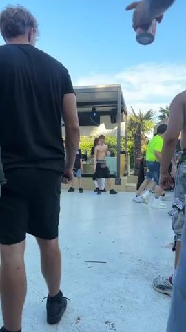 The video shows a group of young people outdoors, possibly at a party or event. The camera is positioned behind some individuals, focusing on their backs and the scene in front of them. The environment appears to be a paved area with some structures and palm trees in the background. The people are casually dressed in t-shirts, shorts, and jeans. Some individuals are standing, while others are in motion. One person in the foreground is wearing a black t-shirt and black shorts, with another person next to them wearing a black t-shirt and denim shorts. In the middle ground, several shirtless men are visible, one wearing camouflage shorts and another in black shorts. A hand with tattoos is seen holding a can, pointing towards the scene. The overall atmosphere seems relaxed and social.