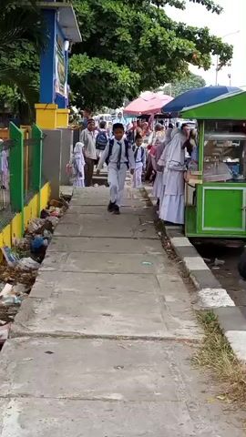 The video captures a street scene with a paved walkway. Several children in school uniforms, including white shirts, green ties, and dark pants or skirts, walk along the walkway. Some carry backpacks. On the left side of the walkway, there is a green metal fence bordering a grassy area with accumulated trash. Behind the fence, a blue and yellow structure is visible, along with trees. On the right side of the walkway, a bright green food cart or stall is present, with people interacting around it. In the background, more people and market stalls are visible under canopies. The weather appears to be daytime and likely warm, given the clothing and outdoor setting. The overall impression is a bustling area with school children navigating a public space with vendors.