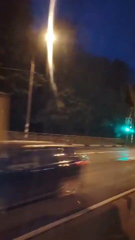 The video shows a car driving on a road at night. The road has multiple lanes and is illuminated by streetlights and the headlights of passing vehicles. There are trees and foliage on the side of the road. A traffic light is visible, showing a green signal. A truck passes by in the opposite direction. Another car with its headlights on is seen in the distance. The overall atmosphere is dark, with the artificial lights creating streaks and reflections due to the motion of the car from which the video is being recorded.