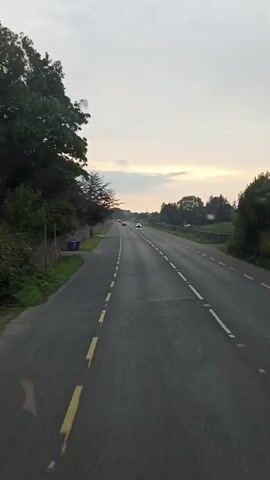 The video shows a straight, paved road with two lanes in each direction, separated by a broken white line. The road is surrounded by green grass and trees. There are stone walls along the side of the road. The sky is cloudy and appears to be dusk. There are several cars visible on the road, driving in both directions. The camera is positioned as if it is inside a moving vehicle, possibly a car or a bus, looking forward. The road markings are yellow and white.