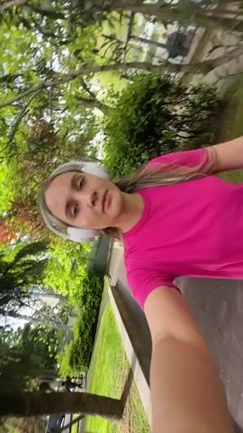 A woman with blonde hair and wearing a pink t-shirt and white headphones walks along a sidewalk. She holds the camera and films herself. Behind her, there are trees and a grassy park area. The sidewalk runs along the edge of the park. Cars and buildings are visible in the background. The day is sunny.